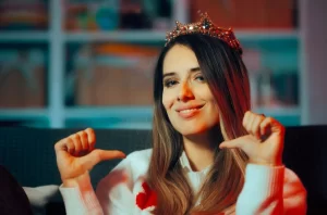 A headshot of a woman wearing a crown, pointing at herself with her thumbs