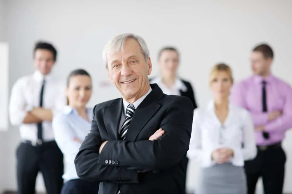 An elderly man in a suit standing with his arms crossed. In the blurry background stand some younger employees